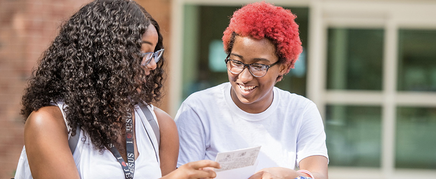 Two female students looking at paper smiling outside on campus.