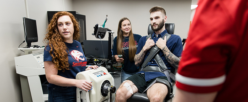 Students working in exercise lab with one student in chair with testing equipment.