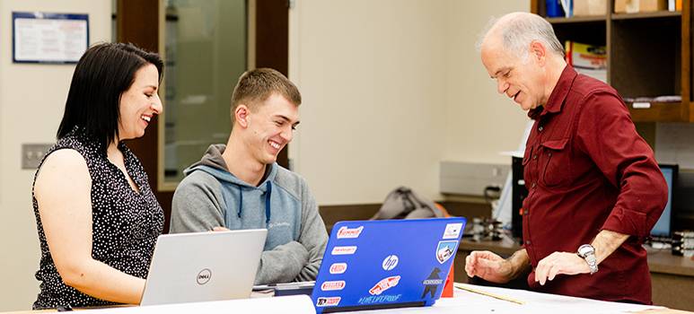 Student smiling looking at laptop with professors looking on.