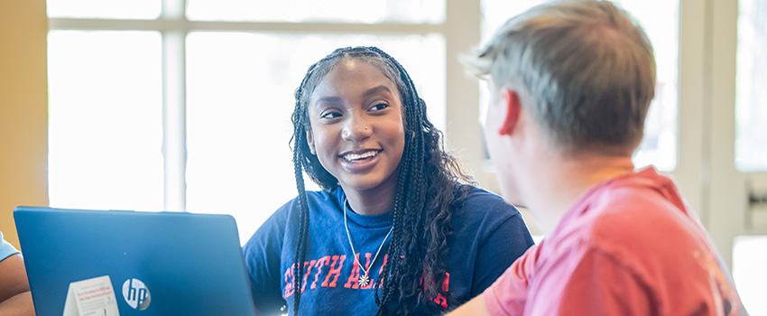 Two students sitting at table with laptop talking.